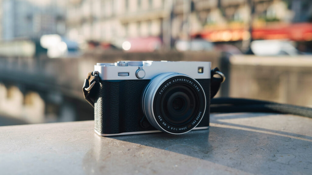 Fujifilm X-E5 argenté sur fond urbain, hybride léger à viseur latéral de style télémétrique, parfait pour la photo de rue et le voyage.